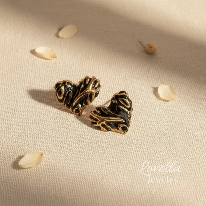 Pair of ornate heart-shaped earrings on a textured beige surface with scattered seeds.