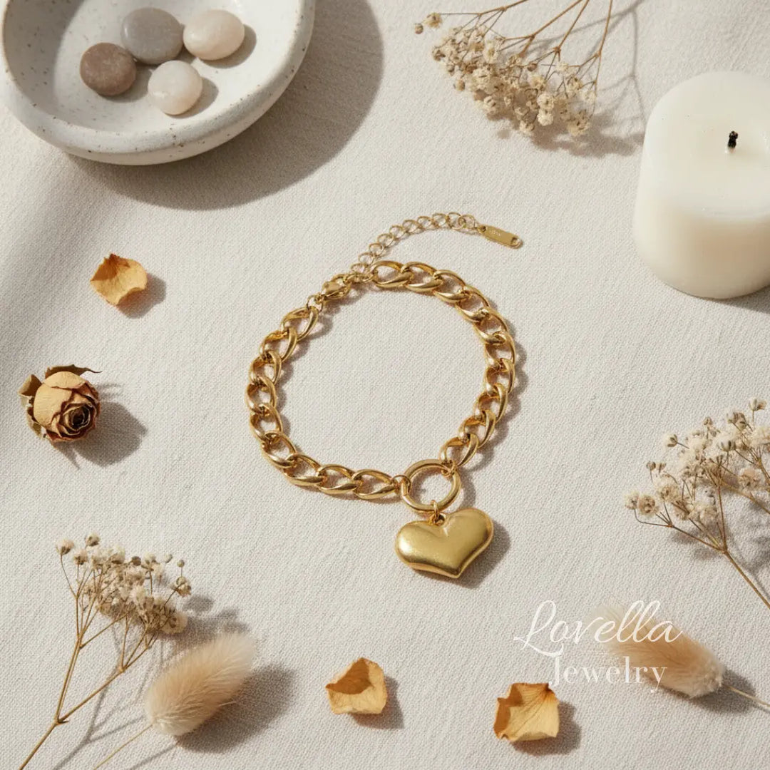 Gold chain bracelet with a heart charm on a light surface with dried flowers and a candle.