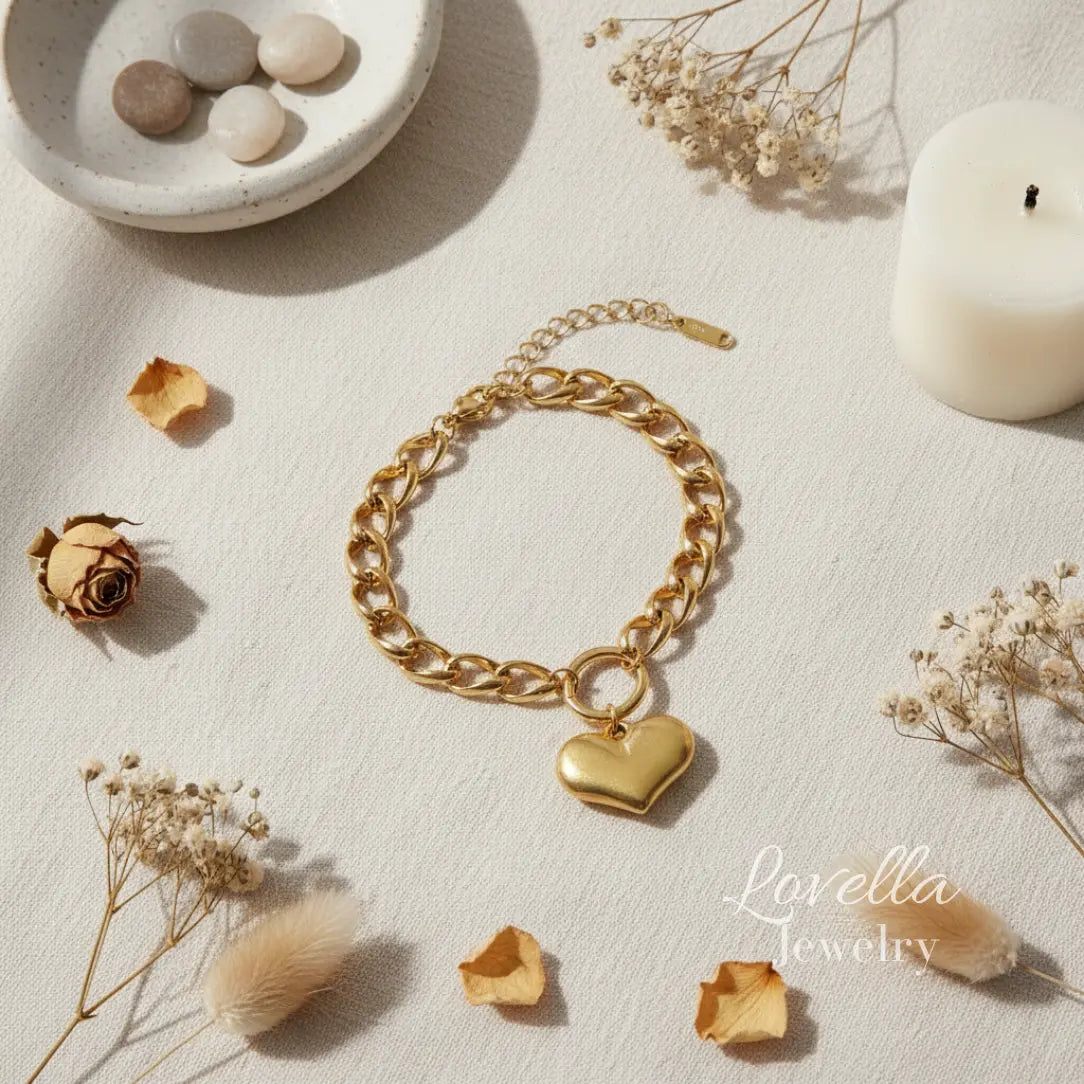 Gold chain bracelet with a heart charm on a light surface with dried flowers and a candle.
