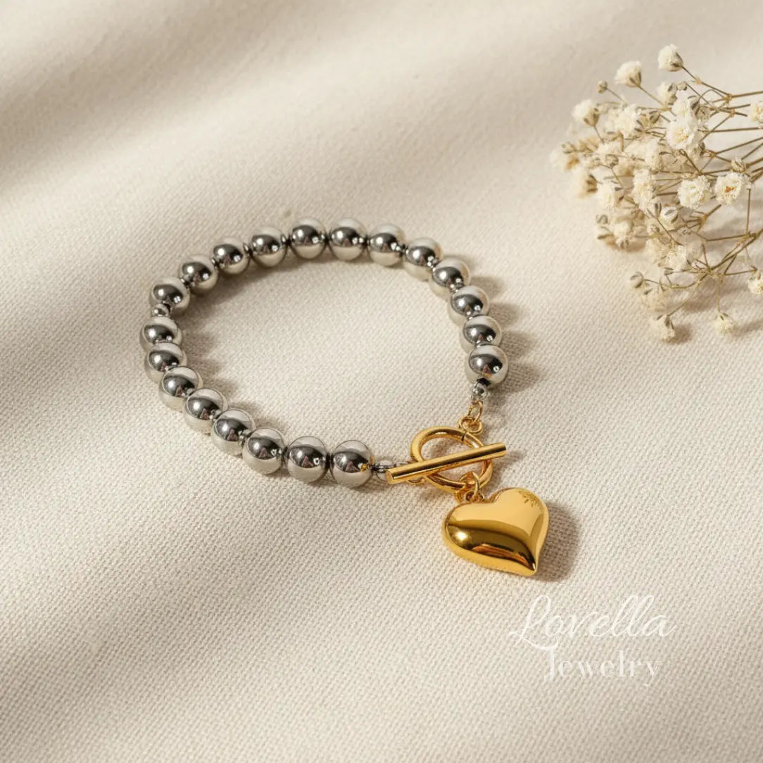 Silver beaded bracelet with a gold heart charm on a beige fabric background