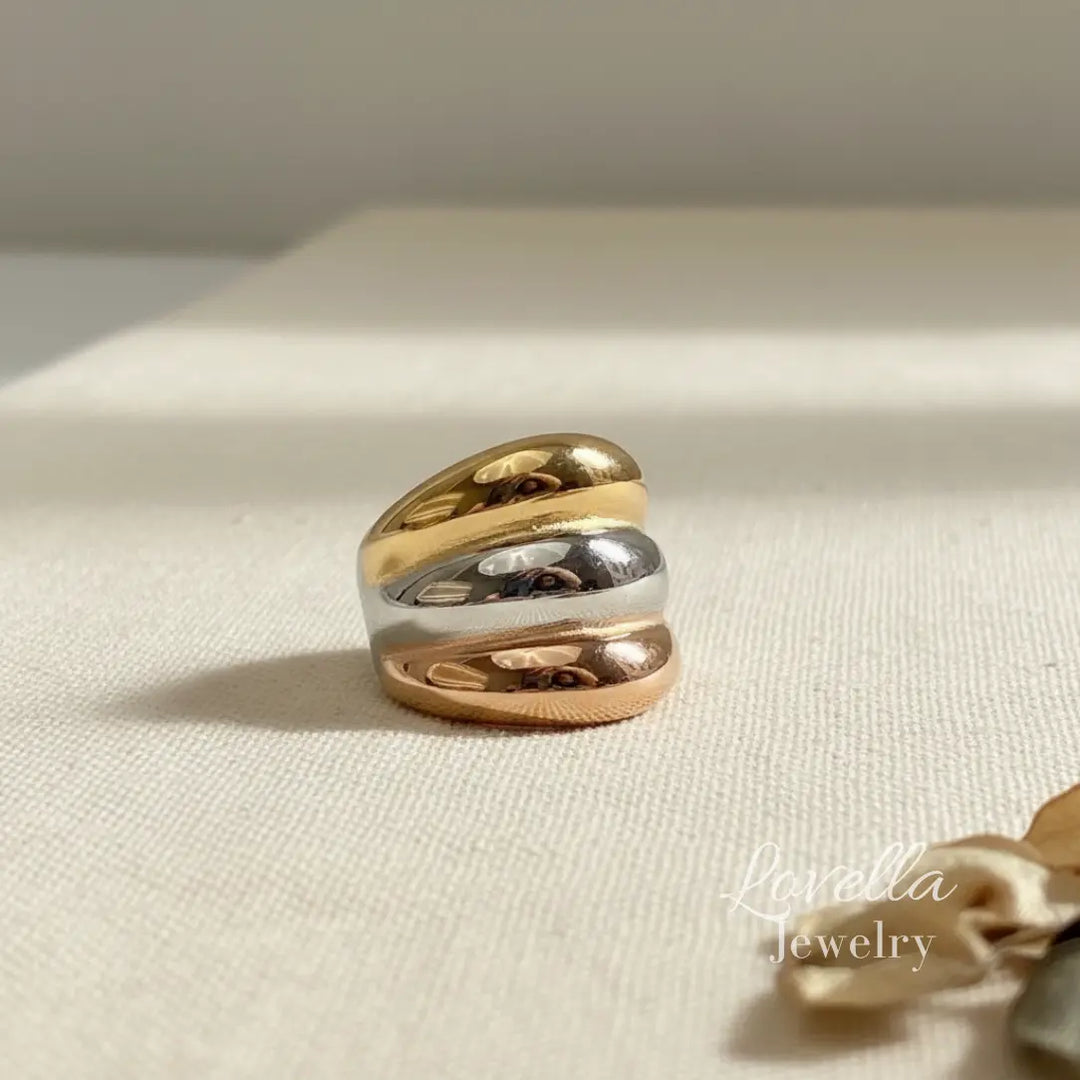 Three-tone chunky band ring with gold, rose gold, and silver layered stripes on cream linen background with dried flowers and warm sunlight, Lovella Jewelry logo.