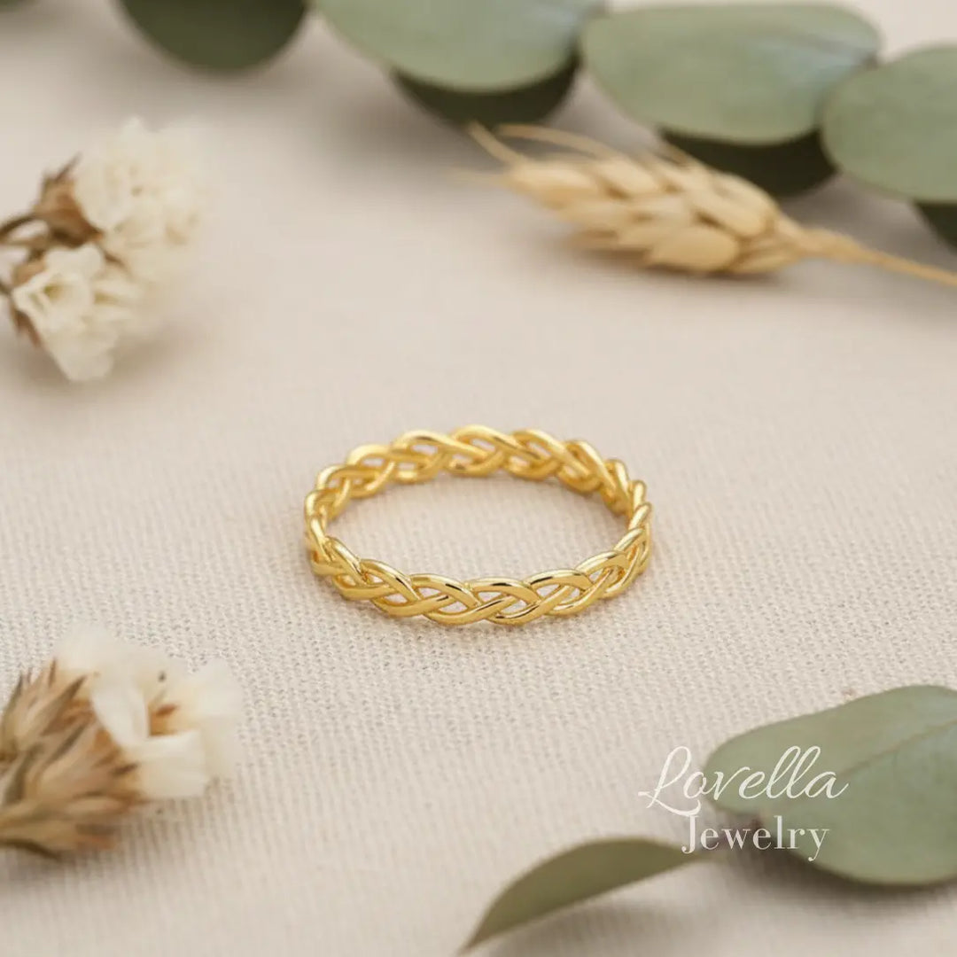Gold-tone stainless steel braided band ring photographed on neutral fabric with soft floral styling.