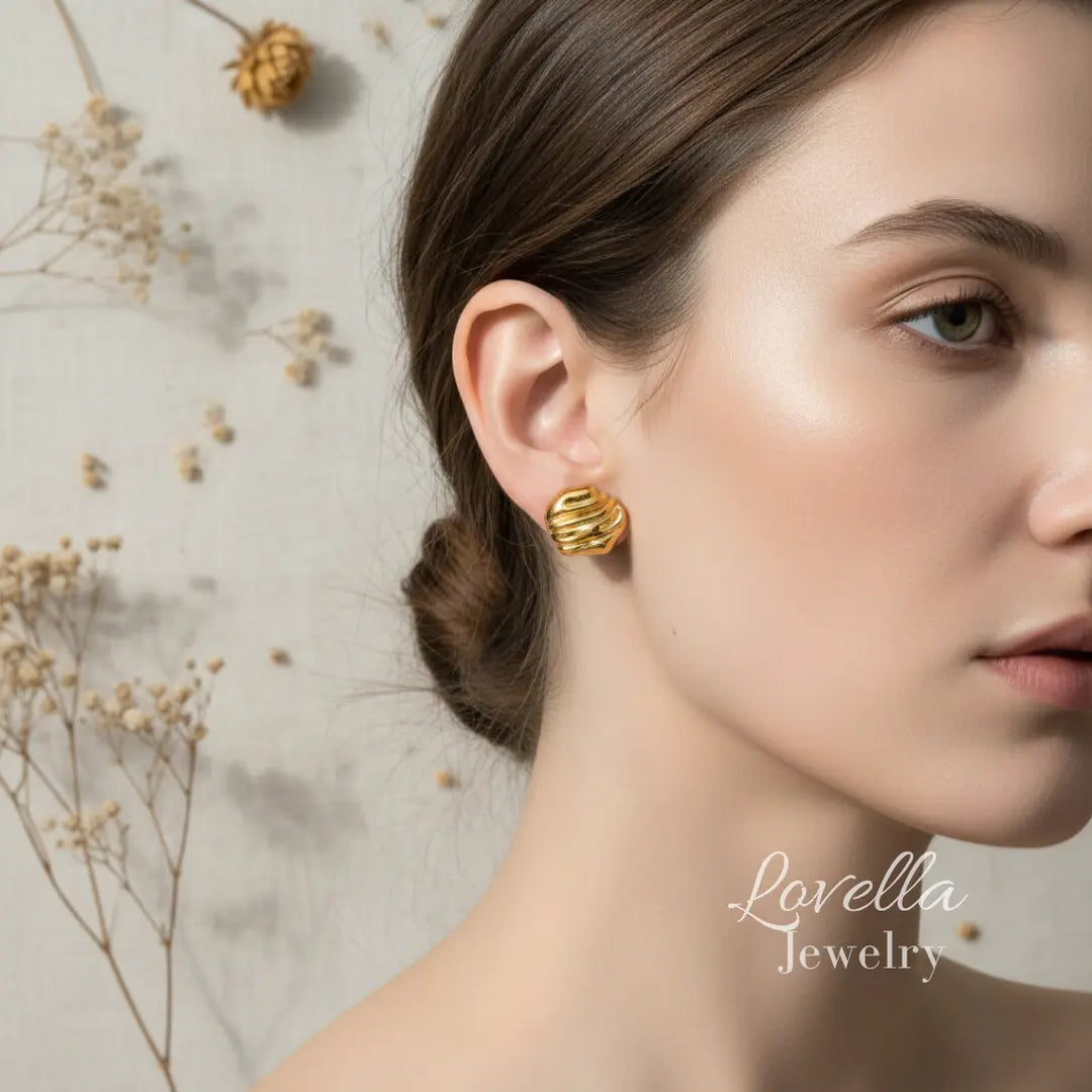 Gold chunky swirl stud earrings worn on ear on cream linen background with dried flowers and Lovella Jewelry logo
