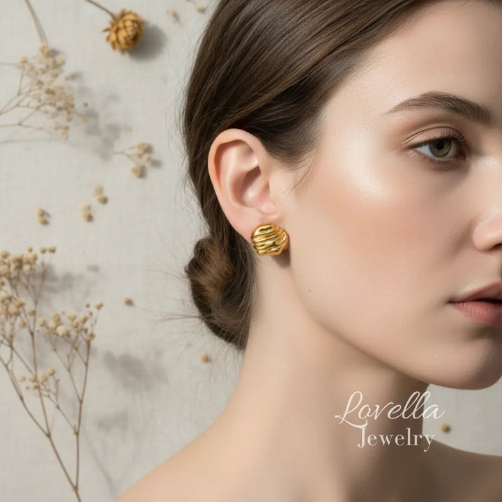 Gold chunky swirl stud earrings worn on ear on cream linen background with dried flowers and Lovella Jewelry logo
