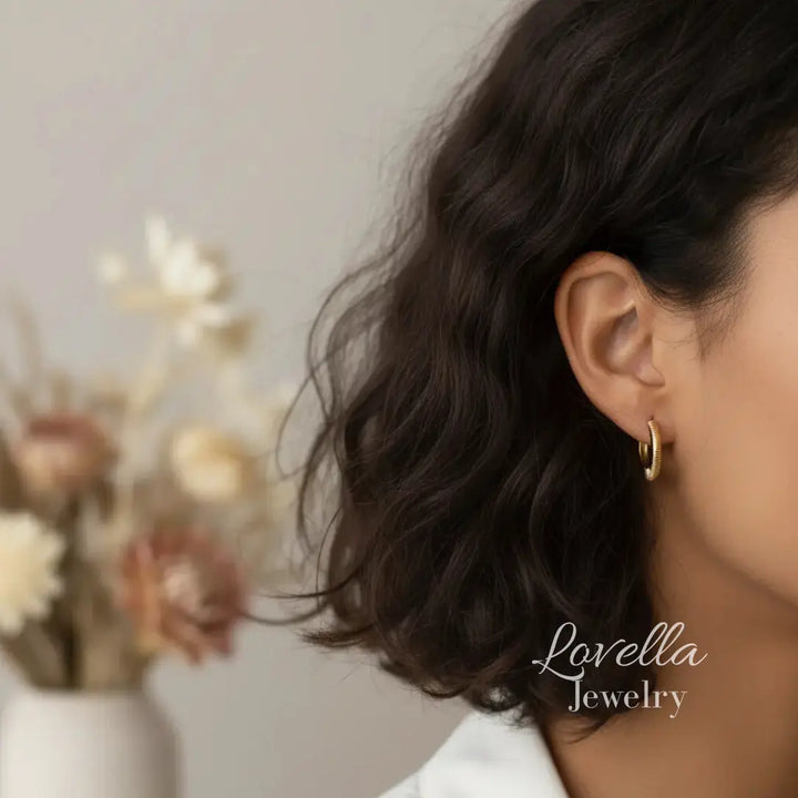 Close-up of a person wearing gold hoop earrings with a blurred background and 'Lovella Jewelry' branding.
