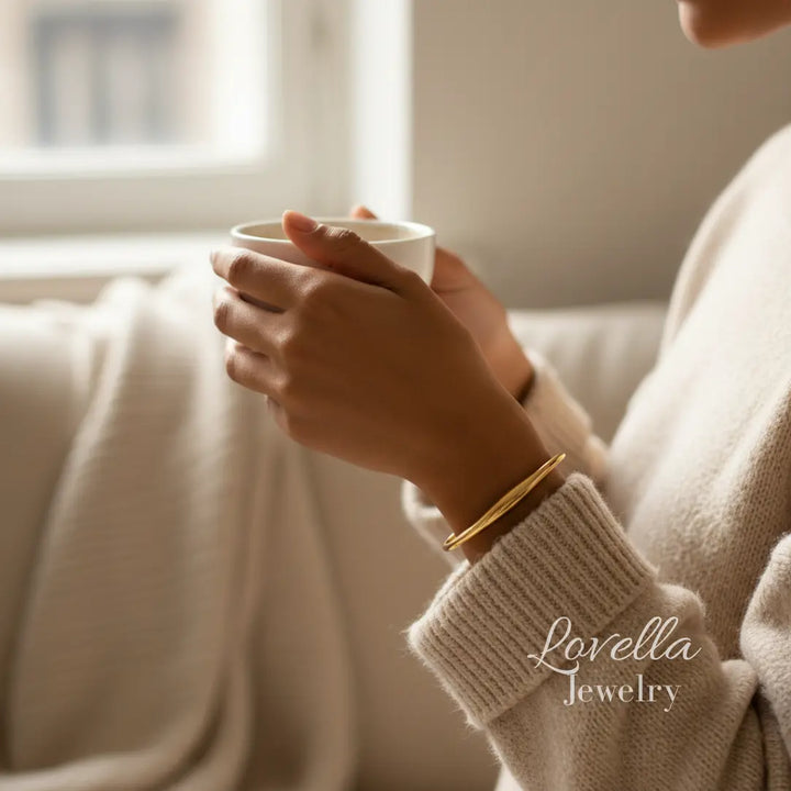 Person holding a white mug with a gold bracelet, wearing a beige sweater, with 'Lovella Jewelry' branding.