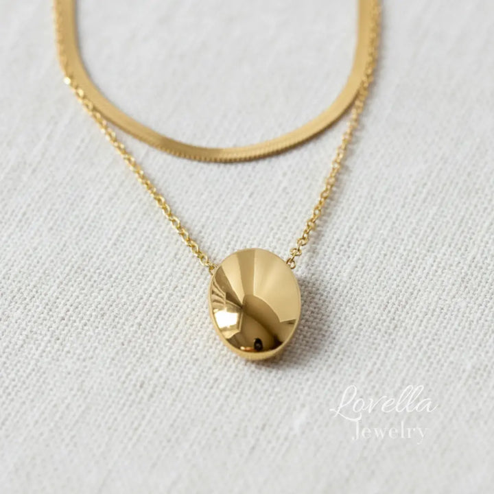 Gold necklace with a round pendant on a white fabric background, featuring Lovella Jewelry branding.