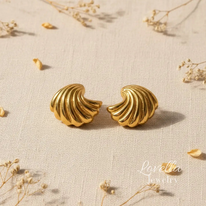 Pair of gold shell-shaped earrings on a textured beige surface with dried flowers.