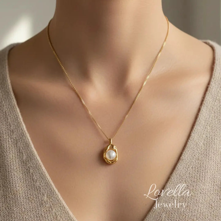 A close-up of a model wearing a delicate gold chain necklace with a teardrop-shaped white pearl pendant by Lovella Jewelry. The model is wearing a beige knit V-neck top, and the background is a soft, neutral gray.