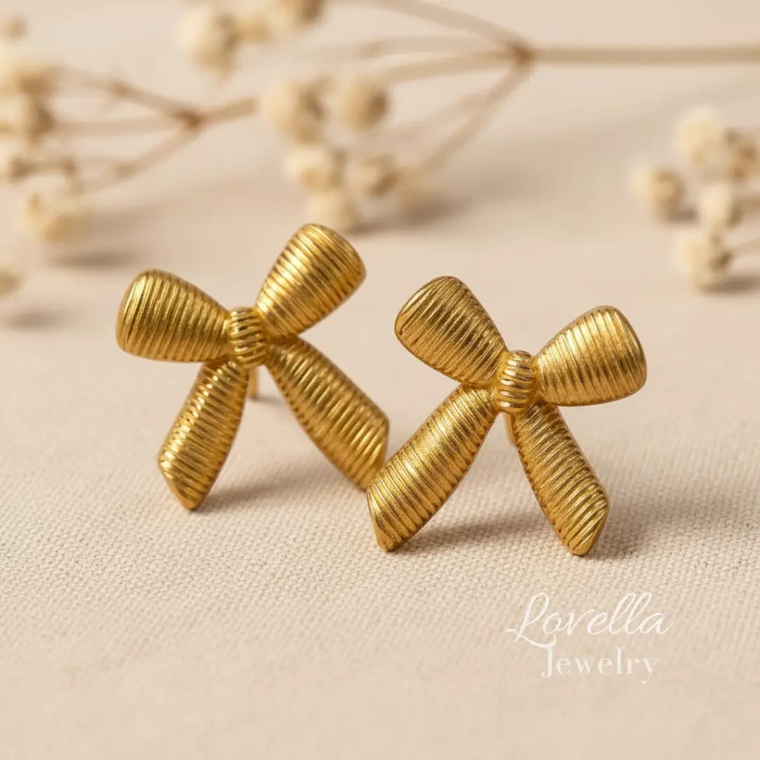 Gold bow-shaped earrings on a beige fabric background with 'Lovella Jewelry' branding.