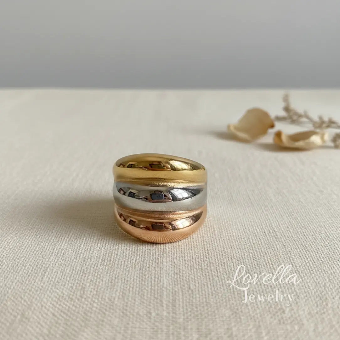 Three-tone chunky band ring with gold, rose gold, and silver layered stripes on cream linen background with dried flowers and warm sunlight, Lovella Jewelry logo.