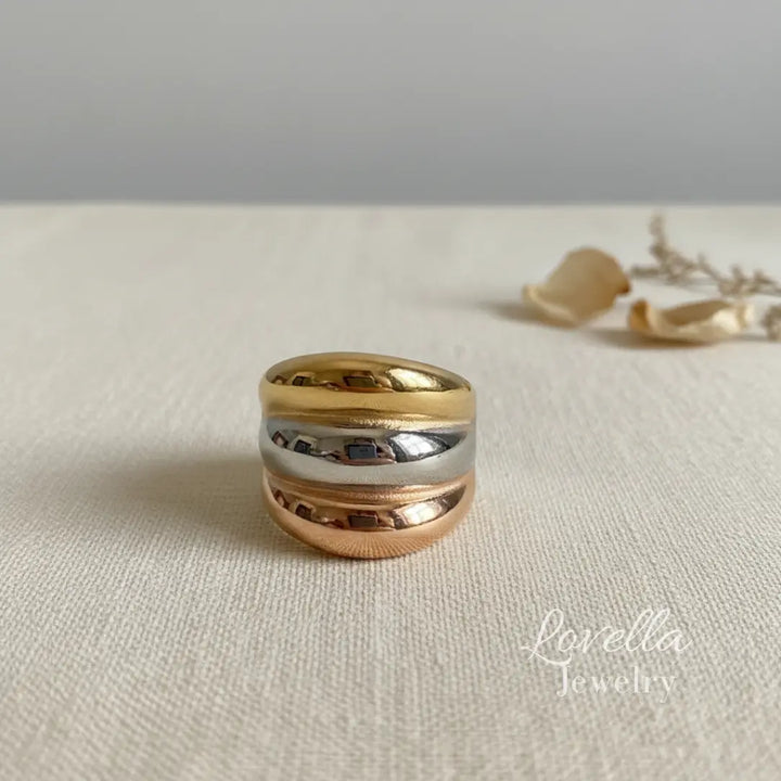 Three-tone chunky band ring with gold, rose gold, and silver layered stripes on cream linen background with dried flowers and warm sunlight, Lovella Jewelry logo.