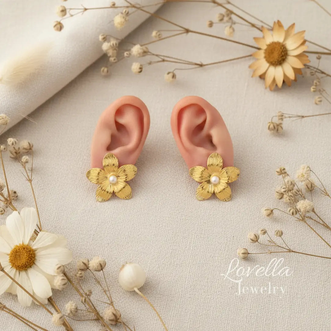 Gold flower earrings with pearl centers on ears surrounded by dried flowers and plants, Lovella Jewelry brand visible.