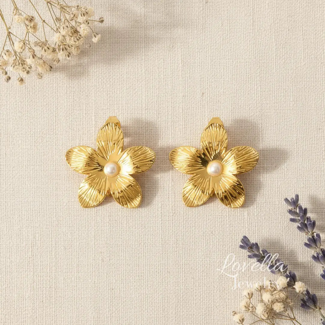 Gold flower-shaped earrings with pearls on a textured beige surface with dried flowers.