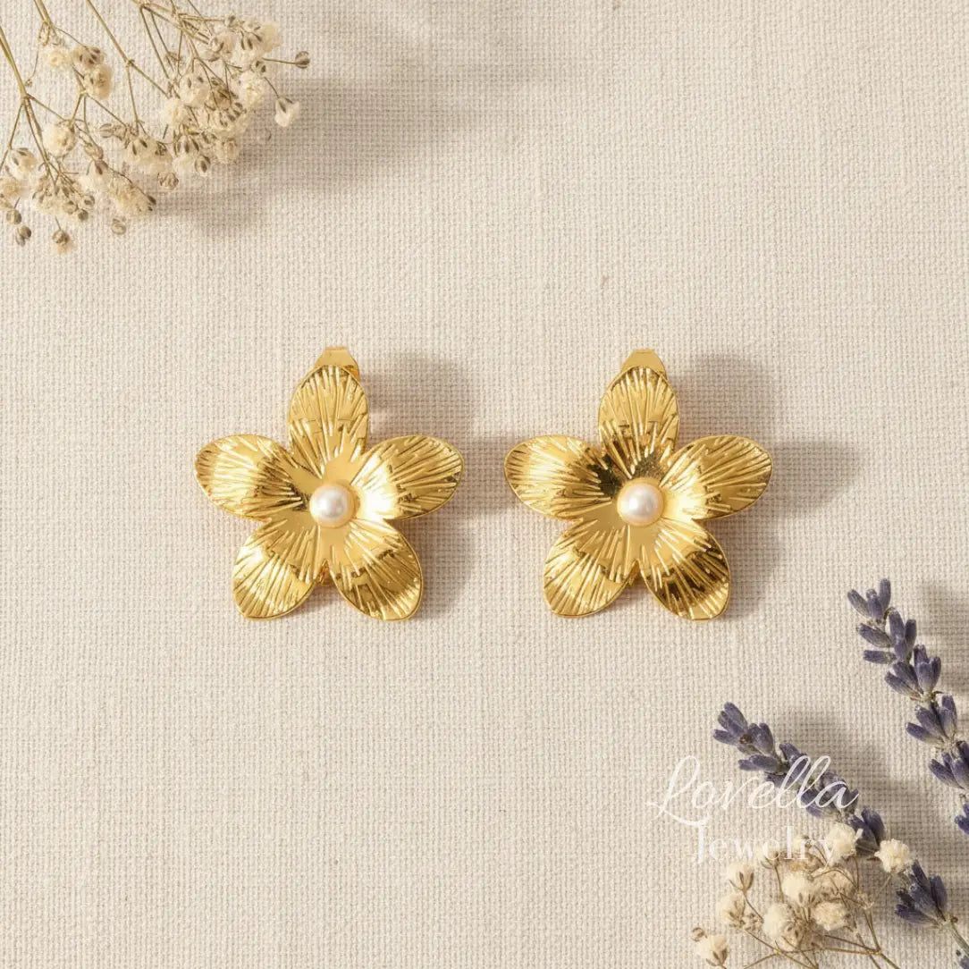 Gold flower-shaped earrings with pearls on a textured beige surface with dried flowers.