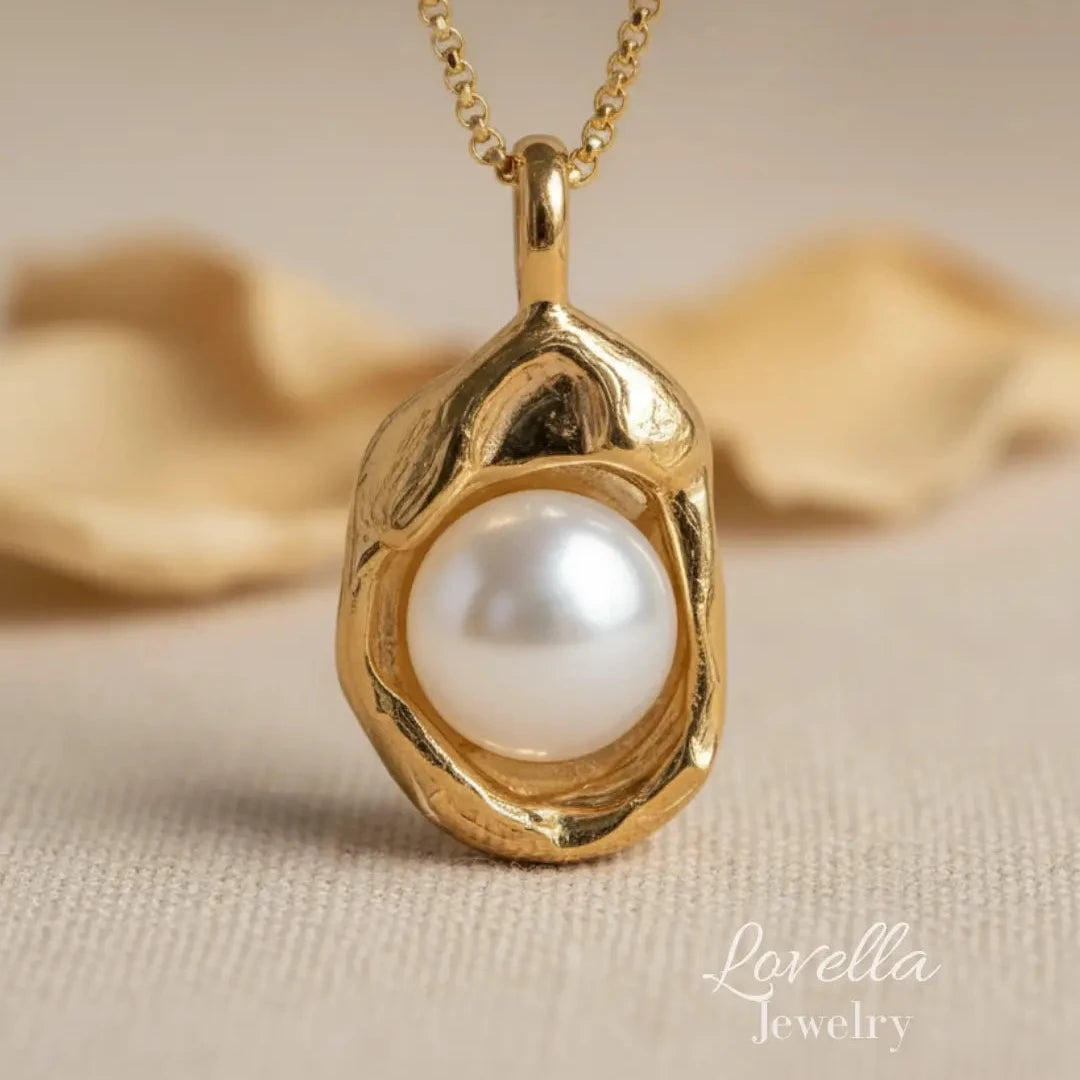 A gold pendant necklace featuring a polished white pearl set in an organic, textured gold bezel, displayed on a soft beige fabric background with a blurred decorative element behind it. The 'Lovella Jewelry' logo is visible in the bottom right corner.