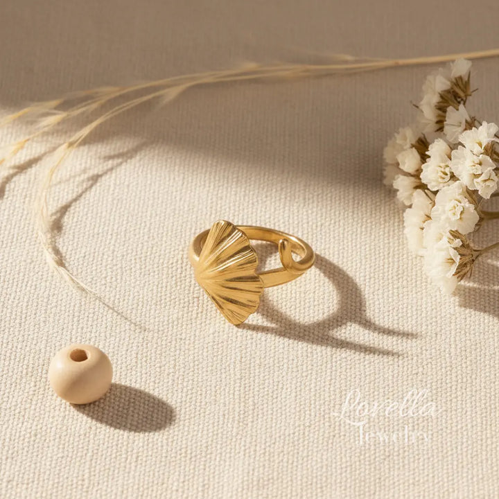 Gold ring with scalloped design on a beige surface with dried flowers and a wooden bead.