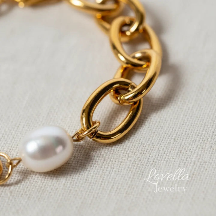 Gold chain bracelet with a pearl on a light fabric background, featuring Lovella Jewelry branding.