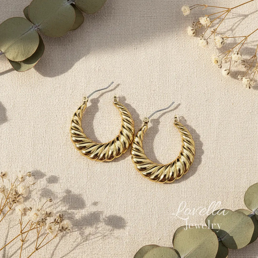 Gold hoop earrings on a textured beige surface with eucalyptus leaves and small flowers.