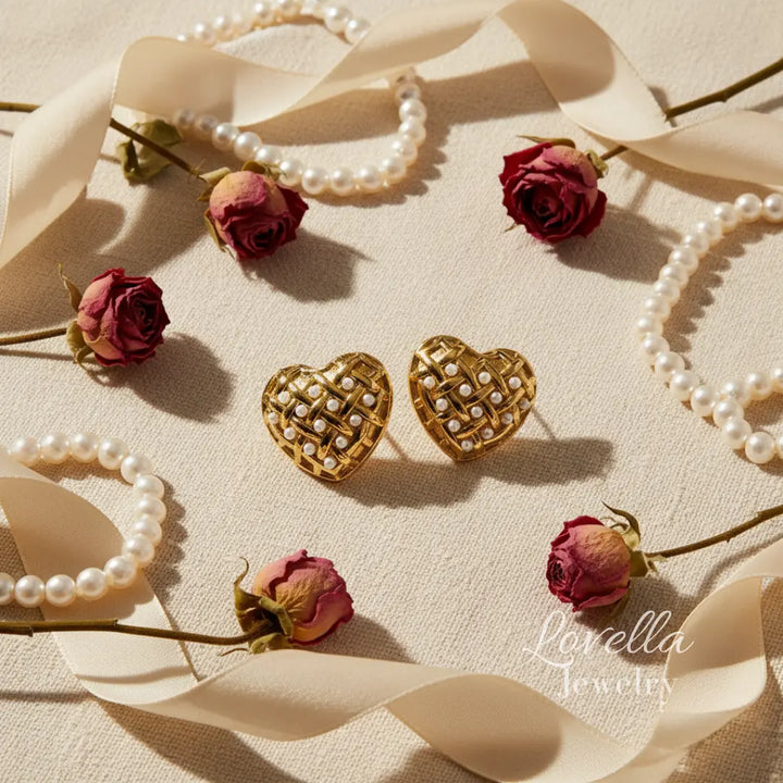 Heart-shaped gold earrings with pearl details on a beige fabric background with pearls and roses.