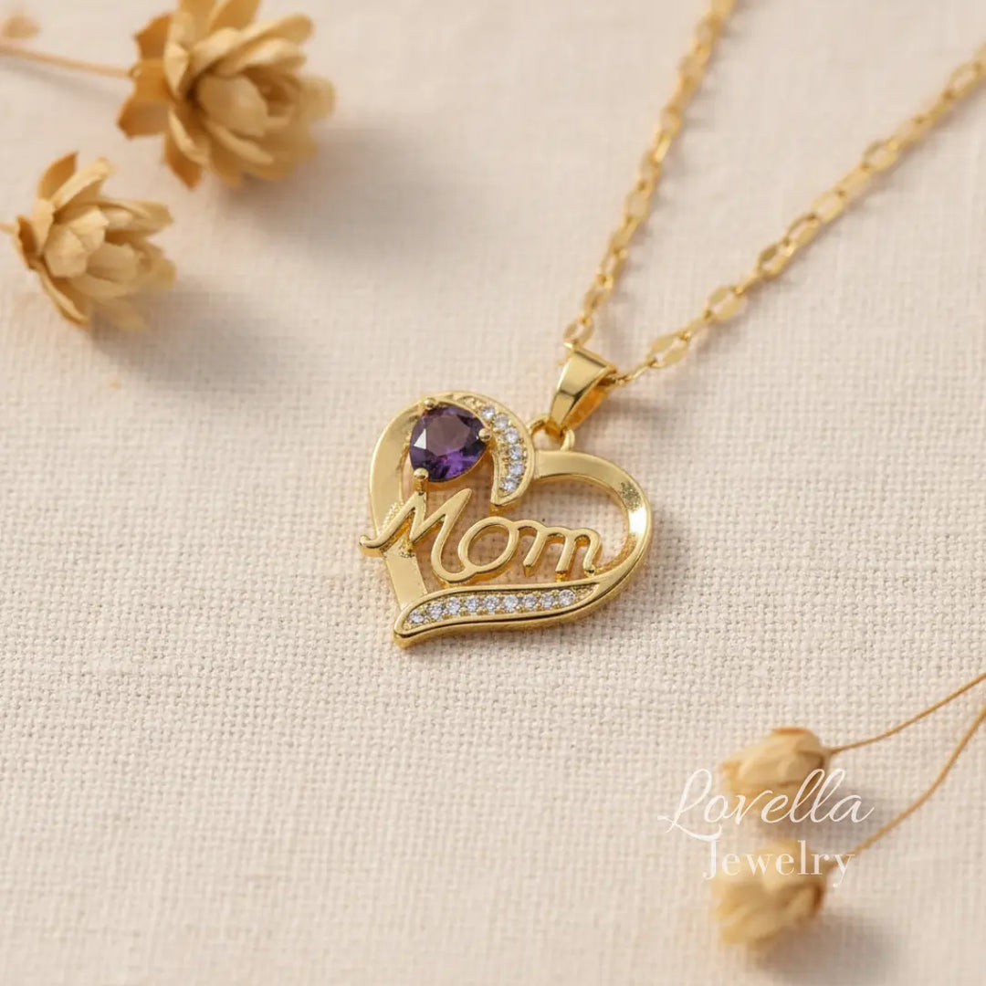 Close-up of gold-tone "Mom" heart pendant necklace featuring a purple center stone and sparkling zirconia details along the heart outline.