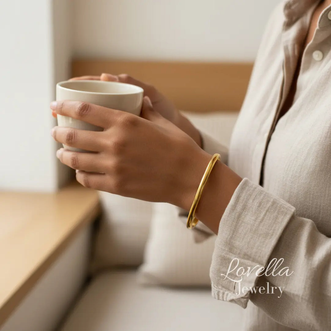 Person holding a mug with a gold bracelet, sitting on a couch with 'Lovella Jewelry' branding.