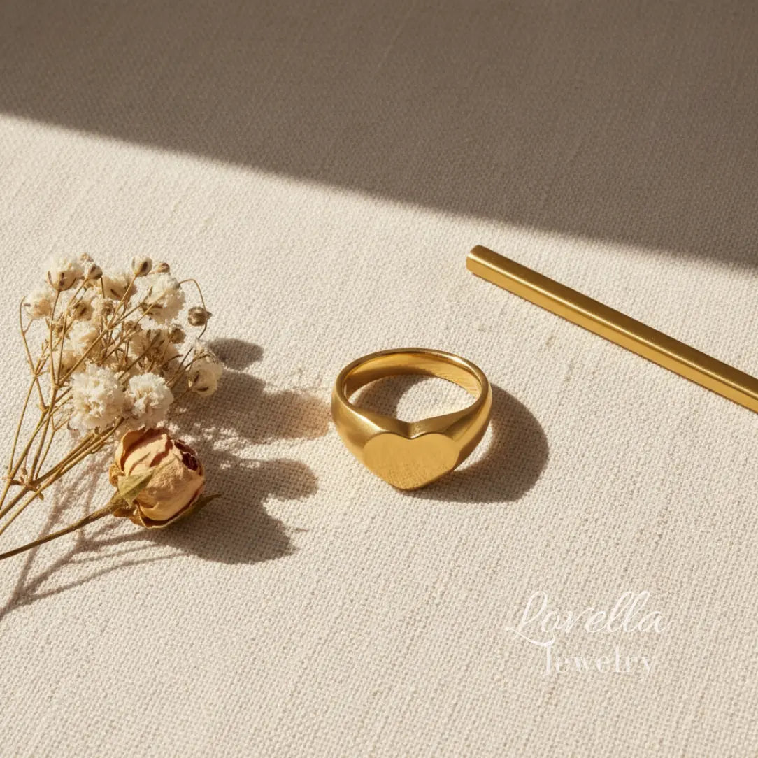 Gold heart-shaped ring on a textured beige surface with dried flowers and a pen.