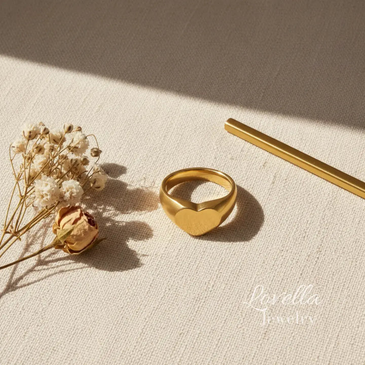 Gold heart-shaped ring on a textured beige surface with dried flowers and a pen.