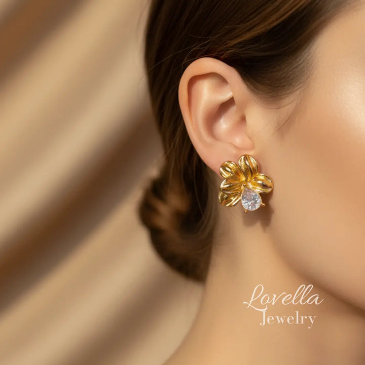 Gold flower-shaped earring worn by a person with a soft beige background, featuring Lovella Jewelry logo.