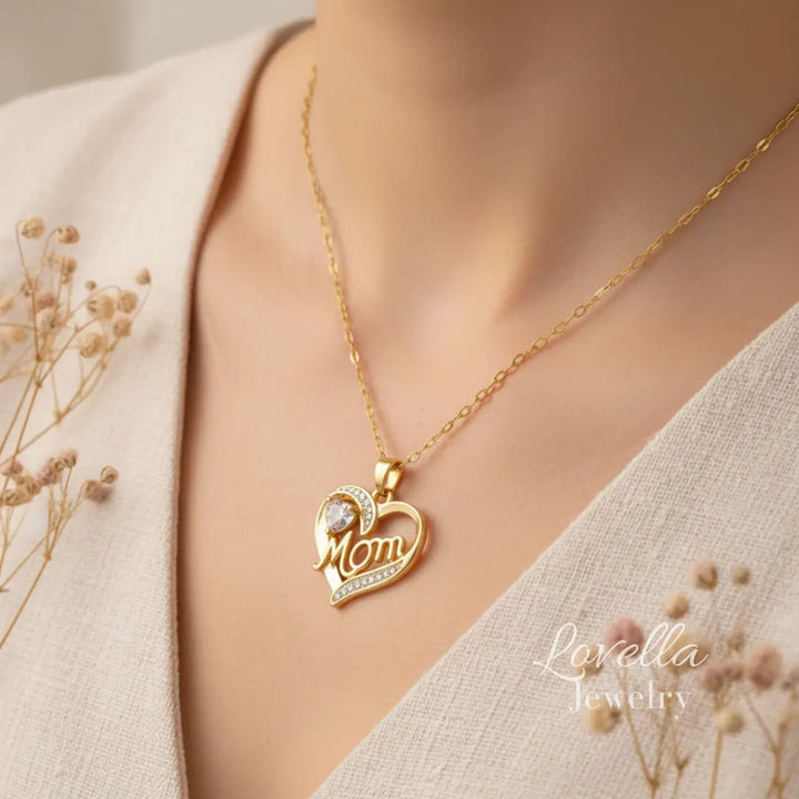 Woman wearing a gold-tone "Mom" heart necklace with white center stone and zirconia accents, styled with neutral tones and dried flowers.