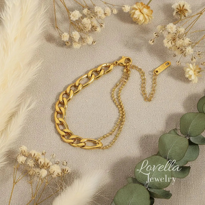 Gold chain bracelet on a textured surface with dried flowers and green leaves, Lovella Jewelry brand visible.