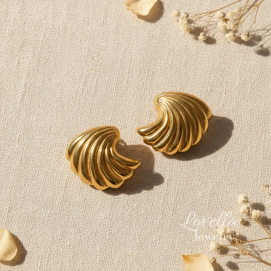 Pair of gold shell-shaped earrings on a textured beige surface with dried flowers.