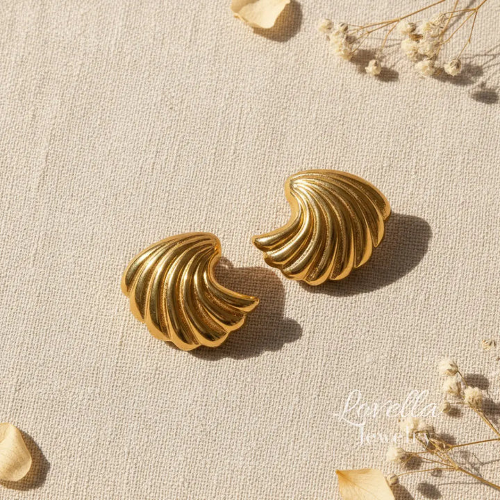 Pair of gold shell-shaped earrings on a textured beige surface with dried flowers.