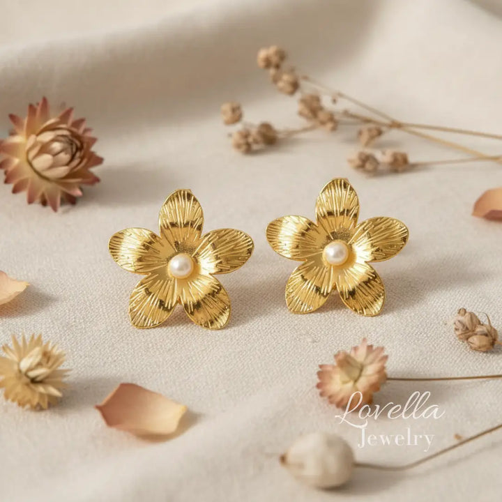 Gold flower-shaped earrings with pearl centers on a beige surface with dried flowers.