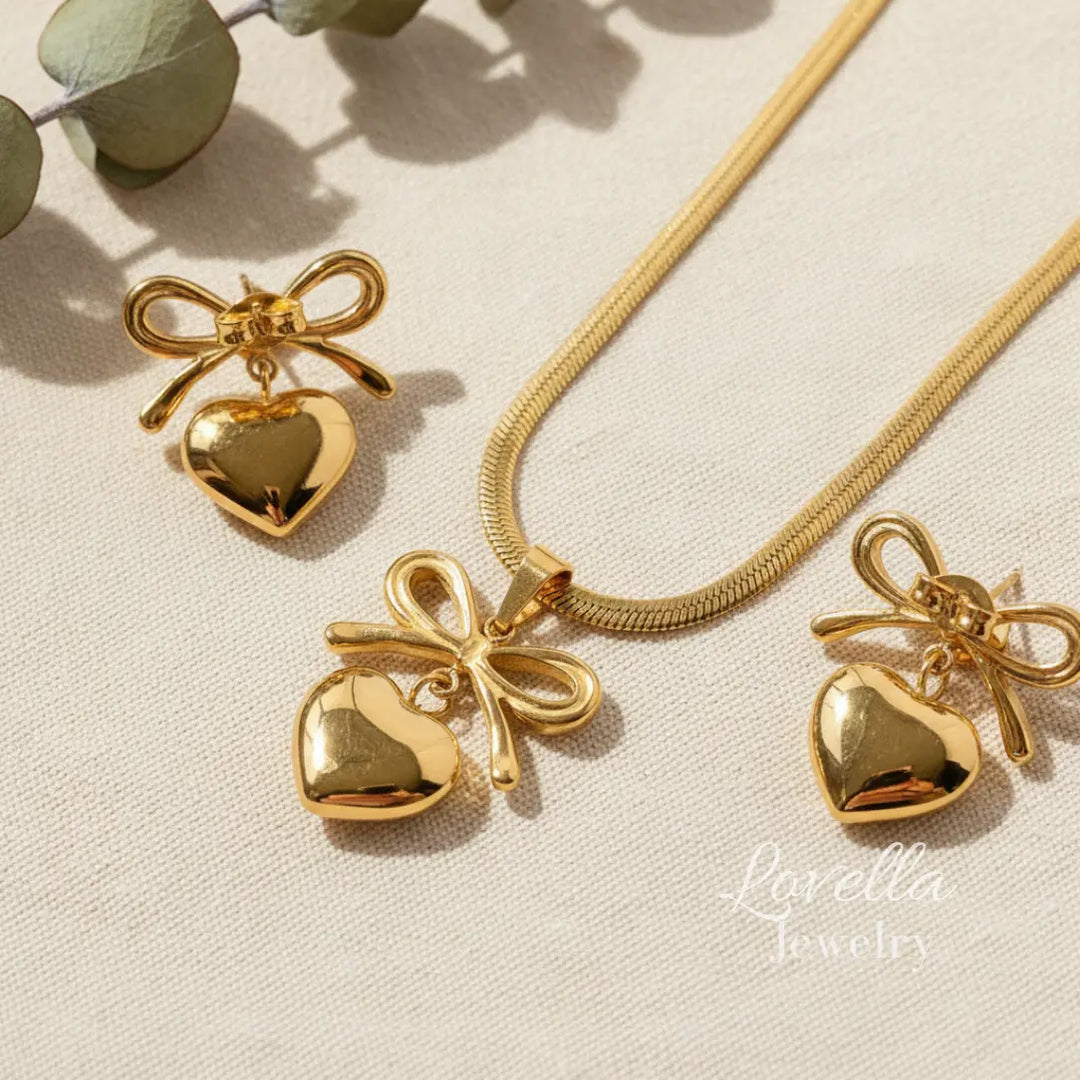 Gold heart-shaped pendants with bows on a beige surface, featuring Lovella Jewels branding.