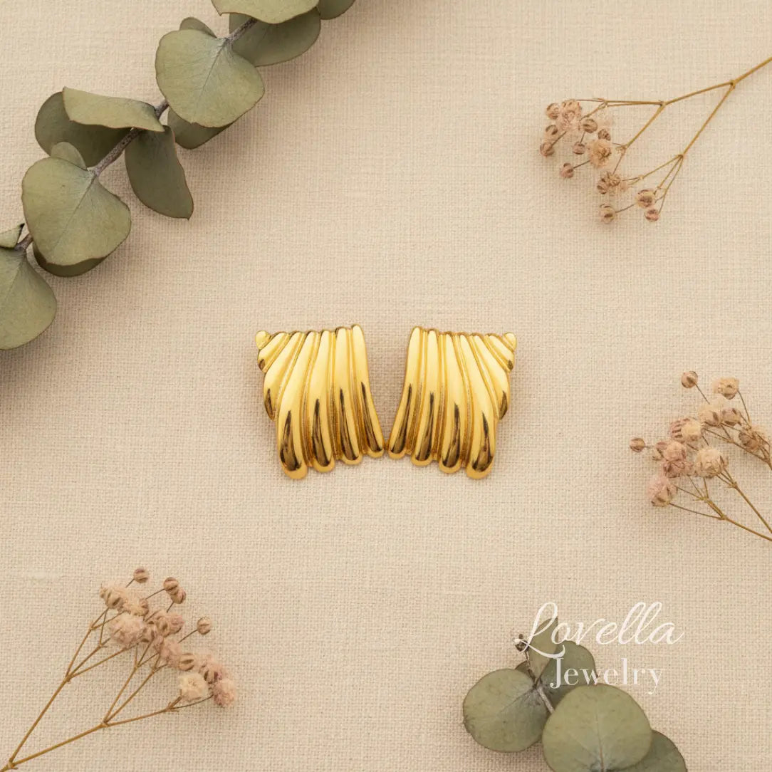 Gold earrings on a beige fabric background with green leaves and dried flowers, Lovella Jewelry branding visible.