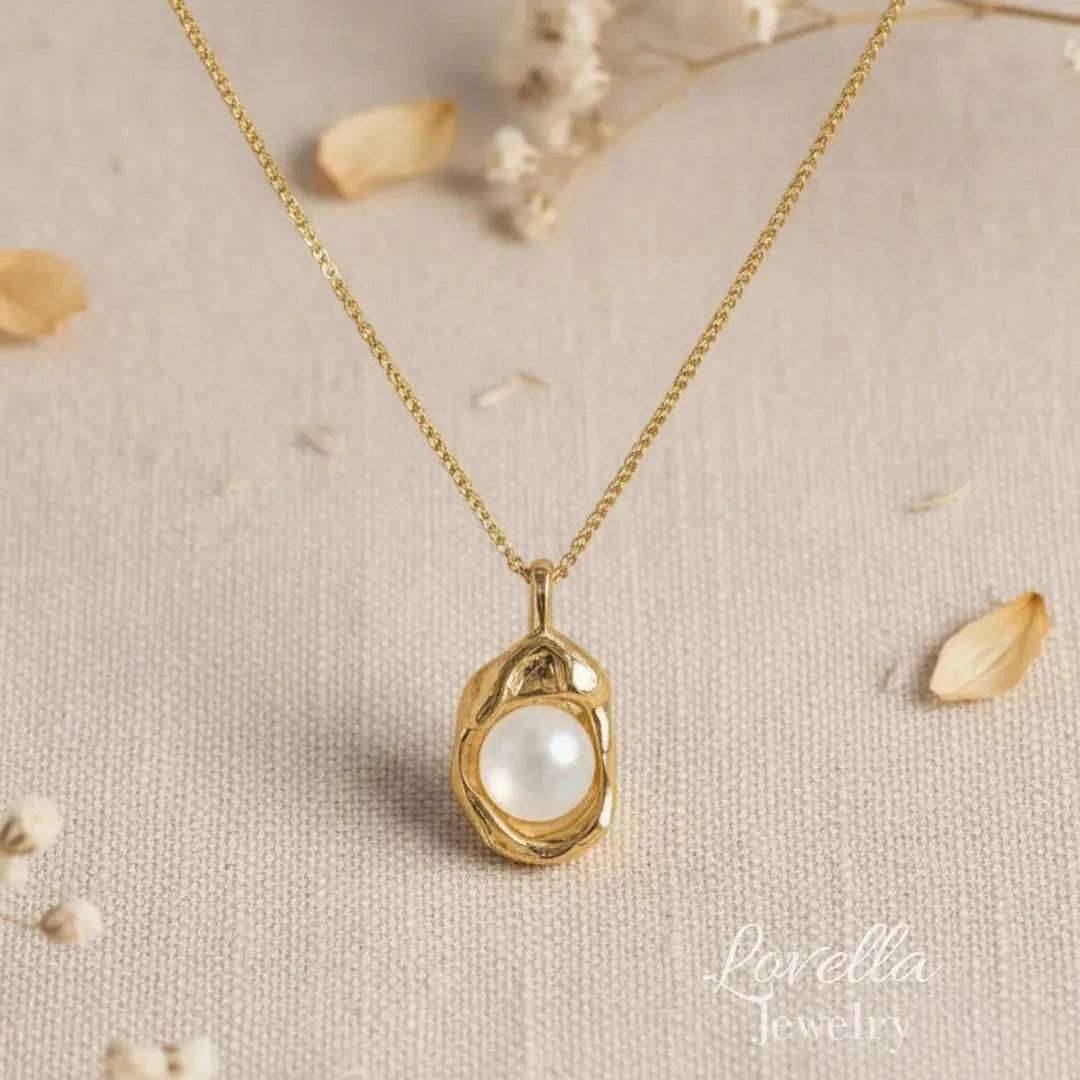 A gold chain necklace with a textured gold pendant holding a white pearl, displayed on a beige fabric background with dried flower petals. The brand name 'Lovella Jewelry' is visible in the bottom right corner.