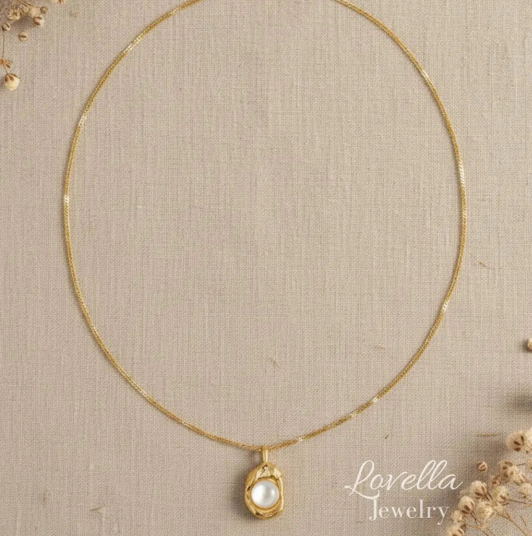 A delicate gold chain necklace with a small oval pendant featuring a white pearl-like center, displayed on a textured beige fabric background with dried floral accents. The brand name 'Lovella Jewelry' is visible in the lower right.