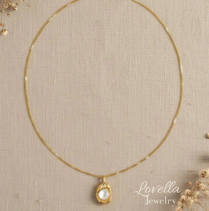 A delicate gold chain necklace with a small oval pendant featuring a white pearl-like center, displayed on a textured beige fabric background with dried floral accents. The brand name 'Lovella Jewelry' is visible in the lower right.