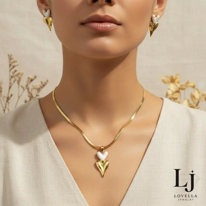 Woman wearing a gold necklace with a heart pendant, earrings, and beige top against a neutral background.