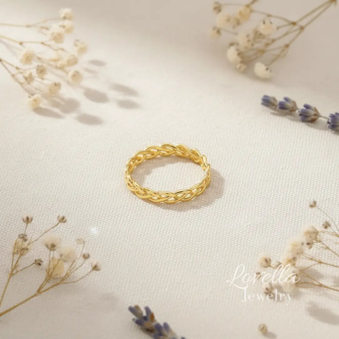 Gold-tone stainless steel braided band ring photographed on neutral fabric with soft floral styling.