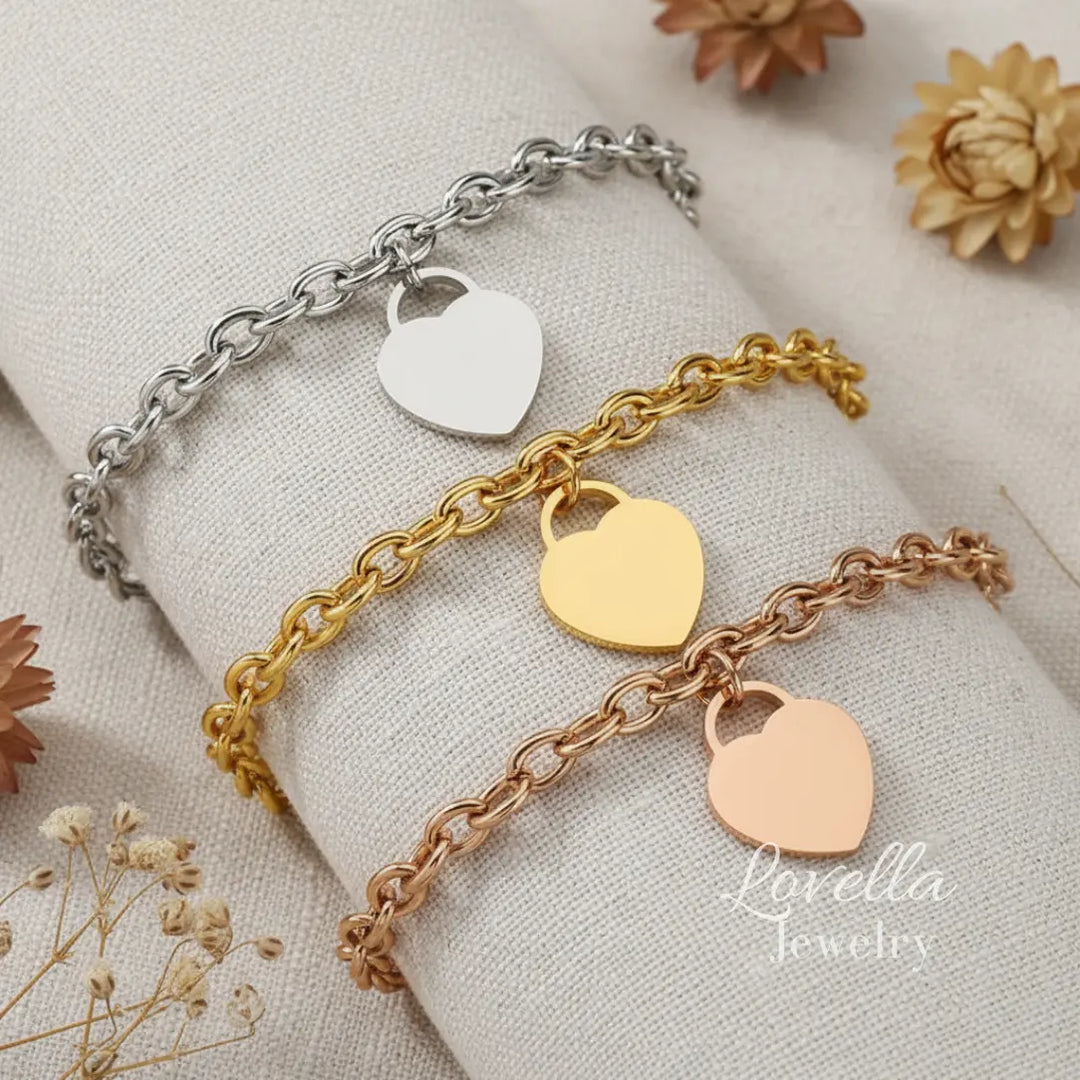 Three heart-shaped charm bracelets in silver, gold, and rose gold on a light fabric background with 'Lovella Jewelry' branding.