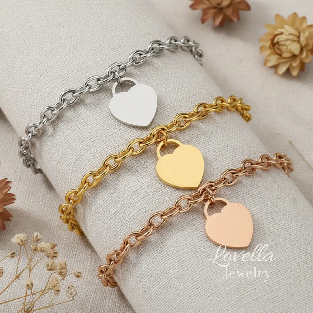 Three heart-shaped charm bracelets in silver, gold, and rose gold on a light fabric background with 'Lovella Jewelry' branding.