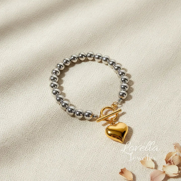 Silver beaded bracelet with a gold toggle clasp and heart charm on a beige fabric background.