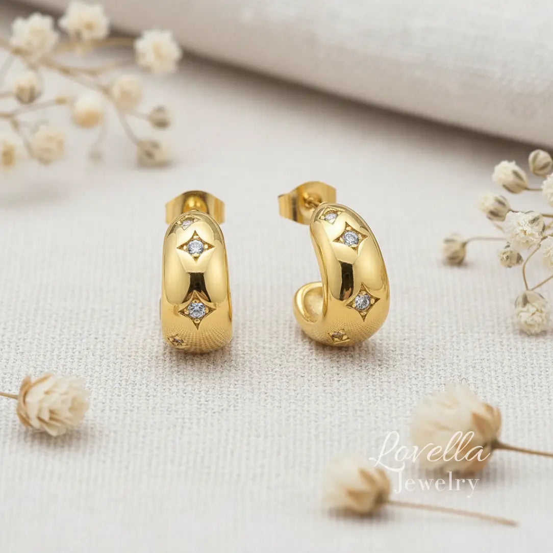 Gold hoop earrings with diamond accents on a textured surface with dried flowers, featuring 'Lovella Jewelry'.