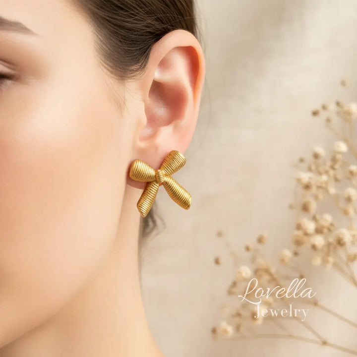Gold bow-shaped earrings worn by a model with a blurred background featuring 'Lovella Jewelry'.