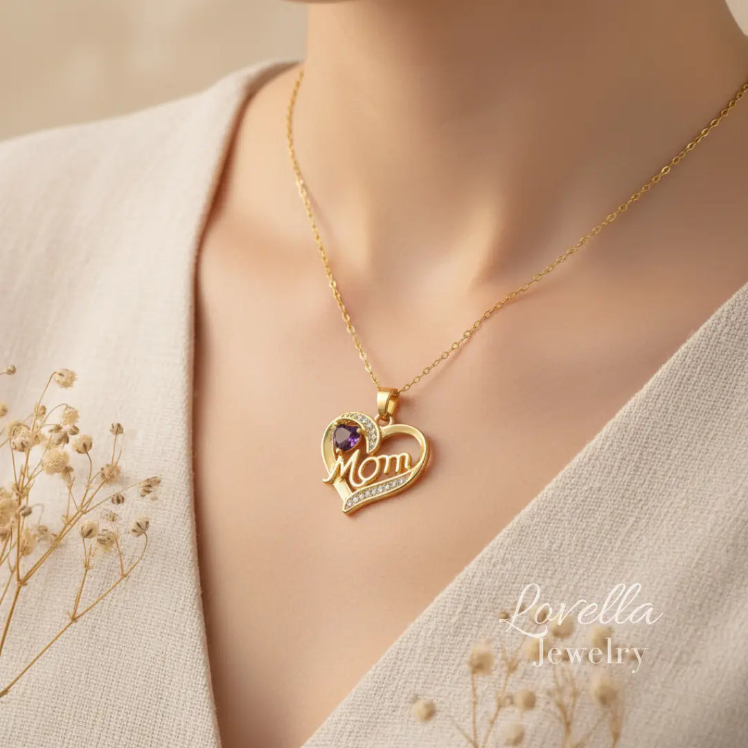Woman wearing a gold-tone "Mom" heart necklace with purple center stone and zirconia accents, styled with neutral tones and dried flowers.