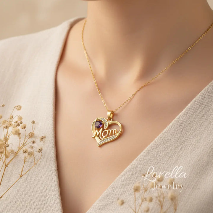 Woman wearing a gold-tone "Mom" heart necklace with purple center stone and zirconia accents, styled with neutral tones and dried flowers.