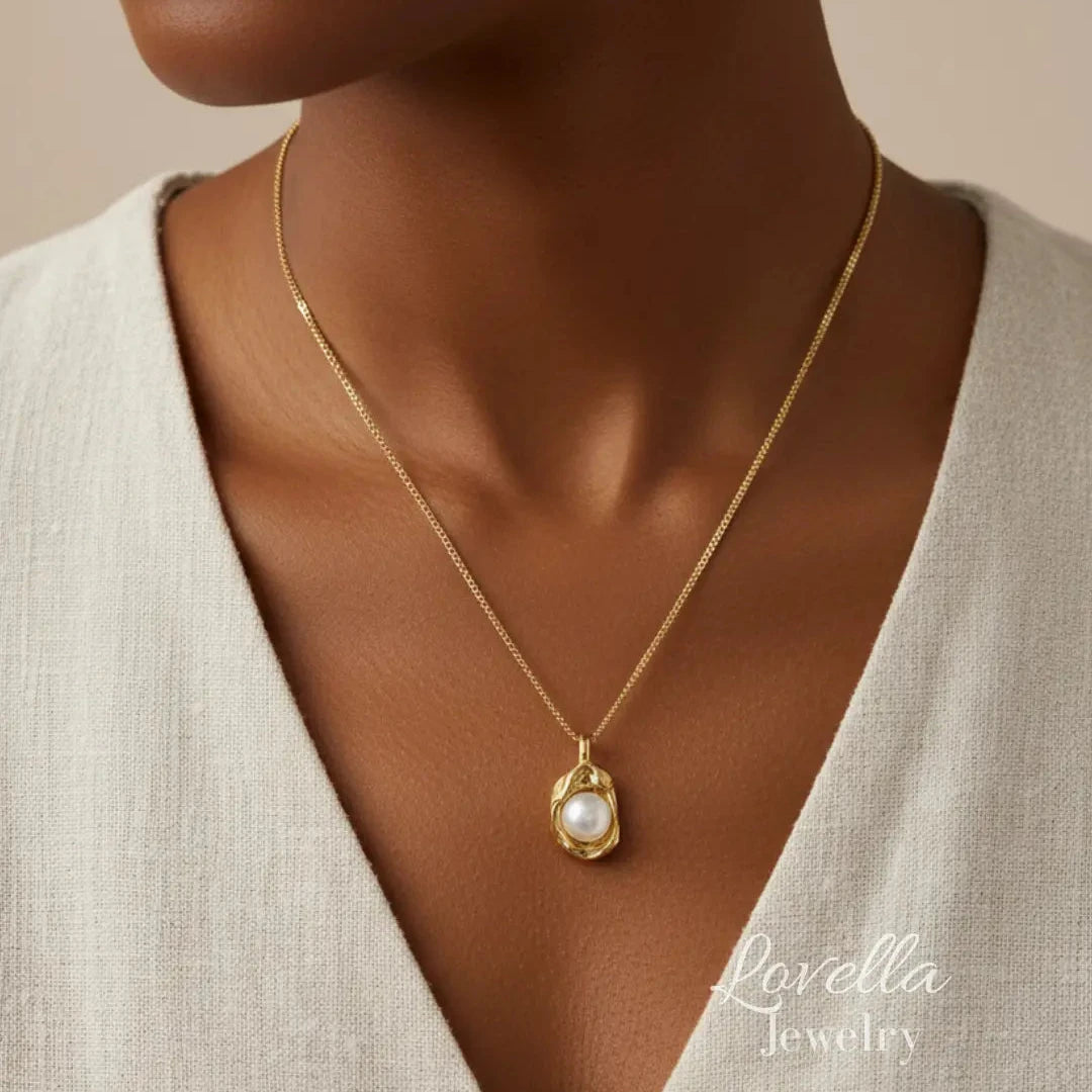 A close-up of a model wearing a delicate gold chain necklace with a teardrop-shaped white pearl pendant. The model is wearing a light beige linen top, and the background is a solid warm beige.