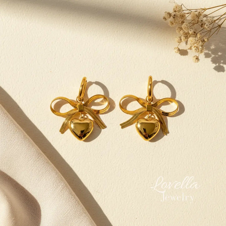 Gold bow-shaped earrings with heart details on a beige surface, featuring Lovella Jewelry branding.